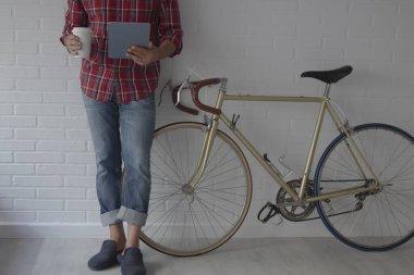 Evdeki adam bisikletin yanındaki duvara yaslanmış tableti kullanıyor ve kahve içiyor.