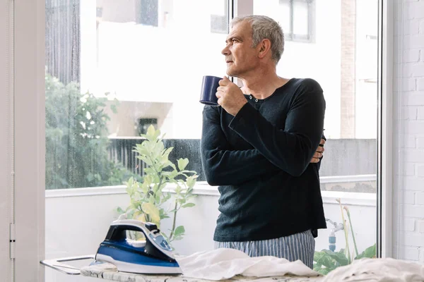 Adam evde gömleğini ütülerken kahve içiyor.