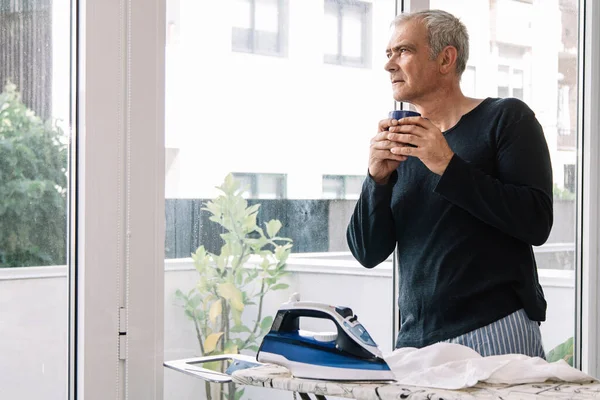 Adam evde gömleğini ütülerken kahve içiyor.