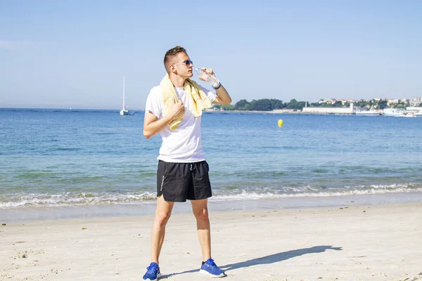 Genç adam plajda dinleniyor ve egzersiz yaptıktan sonra içiyor.