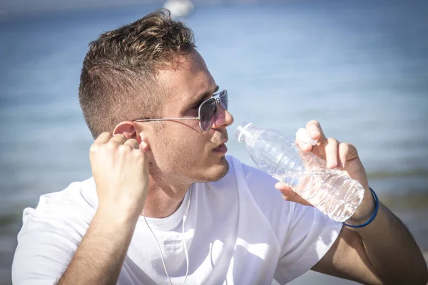 Sahilde dinlenen bir adam spor yaptıktan sonra kulaklıkla ve cep telefonuyla müzik dinliyor.