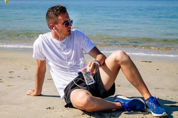 Sahilde dinlenen bir adam spor yaptıktan sonra kulaklıkla ve cep telefonuyla müzik dinliyor.