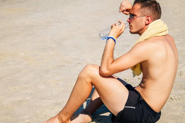Adam egzersiz yaptıktan sonra sahilde dinleniyor ve su içiyor.