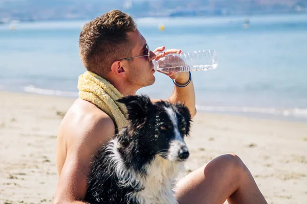 Genç adam sahilde köpeğiyle dinleniyor ve içiyor.