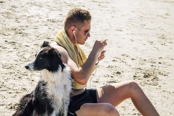 Kumsalda evcil hayvanıyla rahat bir adam cep telefonu kulaklıklarıyla müzik dinliyor.