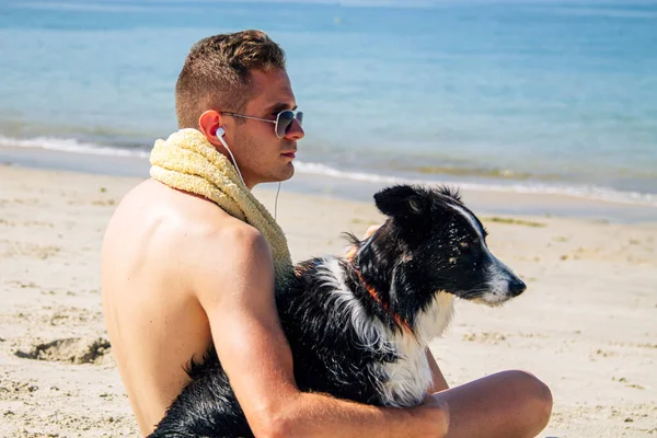 Genç bir adam köpeğiyle sahilde kulaklıkla müzik dinliyor.