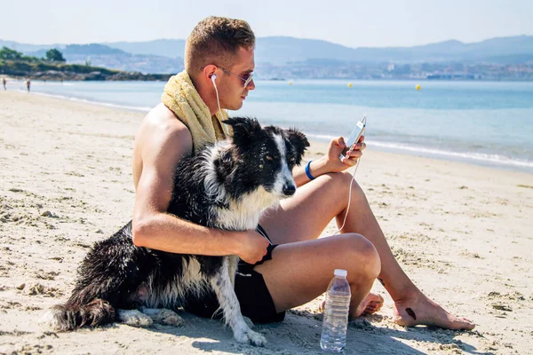 Kumsalda evcil hayvanıyla rahat bir adam cep telefonu kulaklıklarıyla müzik dinliyor.