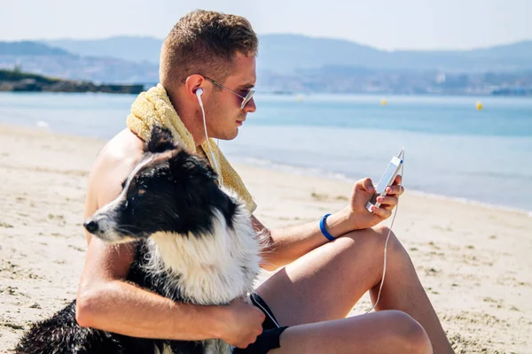 Kumsalda evcil hayvanıyla rahat bir adam cep telefonu kulaklıklarıyla müzik dinliyor.