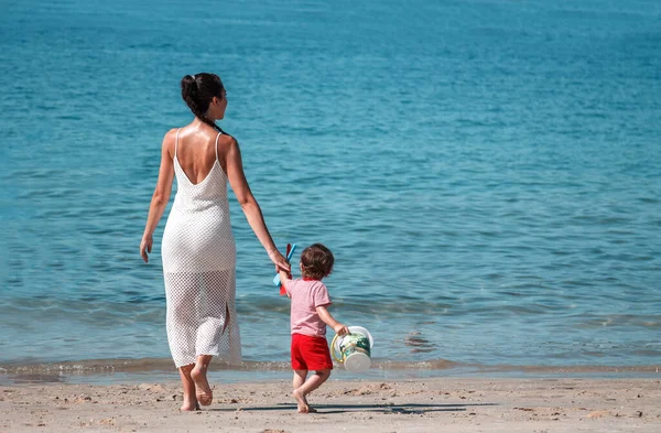 Sırtında çocuğu olan bir kadın sahilde yürüyor.