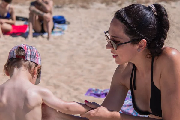 Anne plajdaki çocuğa güneş kremi sürüyor.