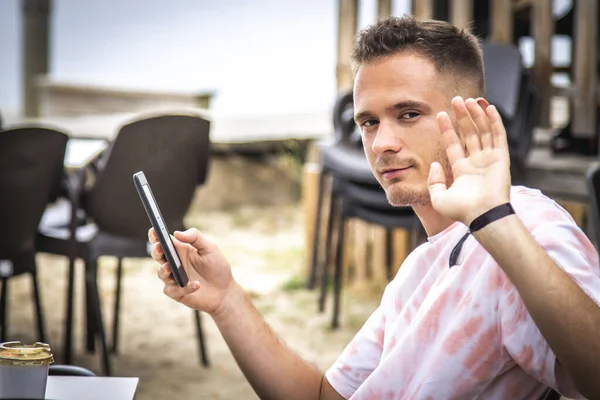 Genç adam cep telefonu kullanıyor ve açık hava bar terasında el sallıyor.