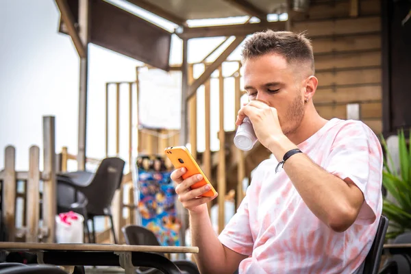 Genç adam cep telefonu kullanıyor ve açık kafede kahve içiyor.