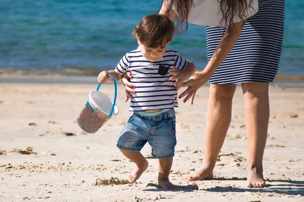 Kumsaldaki küçük çocuk, annesinin elinde kovayla.