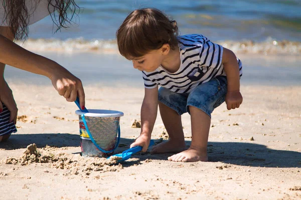 Anne ve küçük oğlu sahilde kova ve kürekle oynuyorlar.
