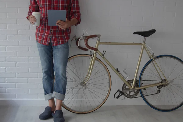 Evdeki adam bisikletin yanındaki duvara yaslanmış tableti kullanıyor ve kahve içiyor.