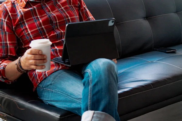 Rahat bir adam kanepede dizüstü bilgisayar kullanıyor ve fotokopi aletiyle kahve içiyor.
