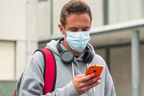 young male student with medical mask and headphones and mobile phone on his way to university or institute