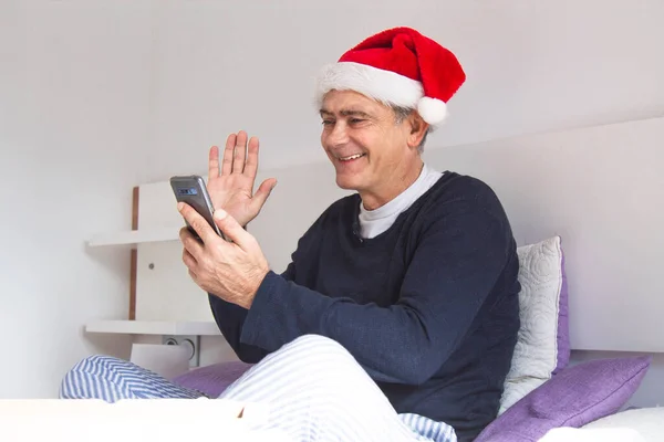 man in pajamas in bed with santa claus hat and mobile phone making video call