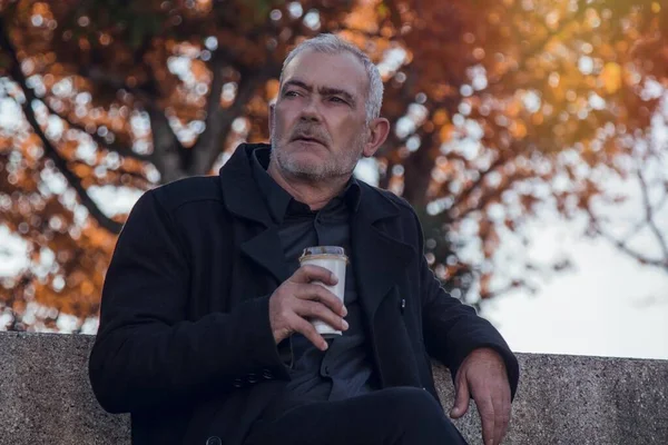 man in coat sitting relaxed on the park bench having coffee