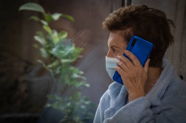 Tıbbi maskeli ve cep telefonlu yaşlı bir kadın pencereden dışarı bakıyor.
