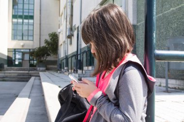 Sırt çantalı öğrenci okulun önündeki cep telefonunu kontrol ediyor.
