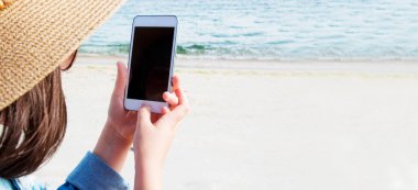 Şapkalı ve kot ceketli kız sahilde cep telefonu kullanıyor.