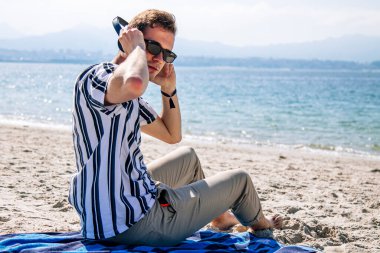 Plajda rahat bir genç adam kulaklıkla müzik dinliyor.