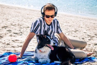 Kumsalda rahatlamış bir adam köpeğiyle eğleniyor ve kulaklıkla müzik dinliyor.