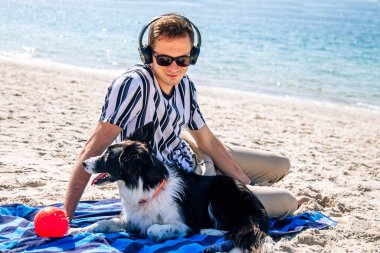 Kumsalda rahatlamış bir adam köpeğiyle eğleniyor ve kulaklıkla müzik dinliyor.