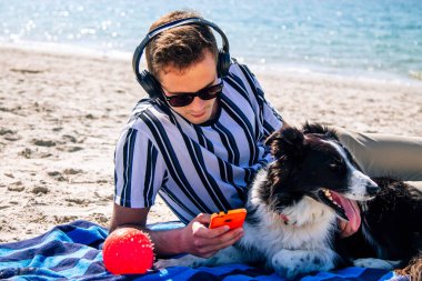 Kulaklıklı ve akıllı telefonlu bir adam kumsaldaki evcil hayvanıyla birlikte.