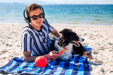 Kulaklıklı ve akıllı telefonlu bir adam kumsaldaki evcil hayvanıyla birlikte.