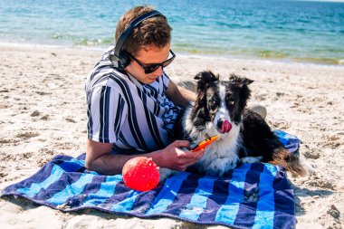 Kulaklıklı ve akıllı telefonlu bir adam kumsaldaki evcil hayvanıyla birlikte.