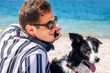 Plajda köpeğiyle cep telefonuyla konuşan genç bir adam.