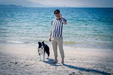 Kumsalda köpeği olan rahat bir adam cep telefonuyla konuşuyor.