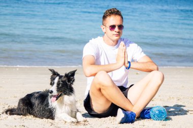 Rahat bir adam köpeğiyle sahilde yoga yapıyor.