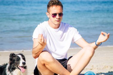 Rahat bir adam köpeğiyle sahilde yoga yapıyor.