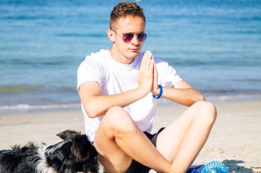 Rahat bir adam köpeğiyle sahilde yoga yapıyor.