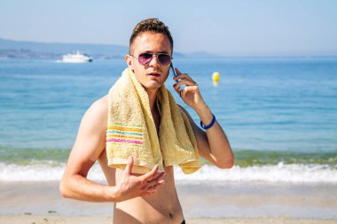 Kumsalda gülümseyen genç adam cep telefonuyla konuşuyor.