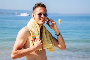 Kumsalda gülümseyen genç adam cep telefonuyla konuşuyor.