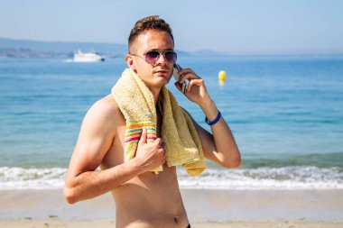 Kumsalda gülümseyen genç adam cep telefonuyla konuşuyor.