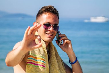 Genç adam plajda cep telefonuyla yürüyor ve parmaklarıyla 