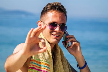 Genç adam plajda cep telefonuyla yürüyor ve parmaklarıyla 