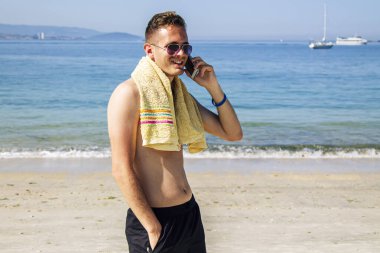 Genç adam plajda cep telefonuyla yürüyor.