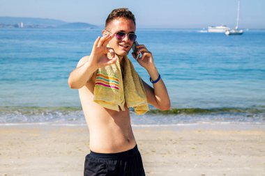 Genç adam plajda cep telefonuyla yürüyor ve parmaklarıyla 