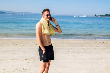 Genç adam plajda cep telefonuyla yürüyor.