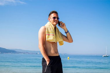 Genç adam plajda cep telefonuyla yürüyor.