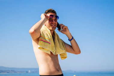 Sahilde güneş gözlüklü ve cep telefonlu genç bir adam.