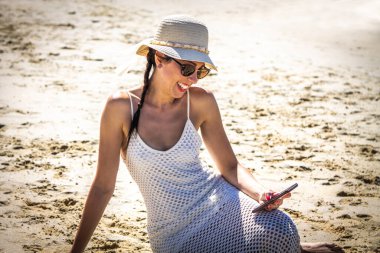 Kumsalda gülümseyen ve rahat bir kadın akıllı telefon kullanıyor.