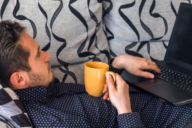 Rahatlamış genç adam dizüstü bilgisayarında yazarken kahve içiyor.