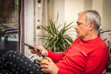 Rahat yetişkin bir adam akıllı telefon kullanıyor ve evdeki kanepede kahve içiyor.